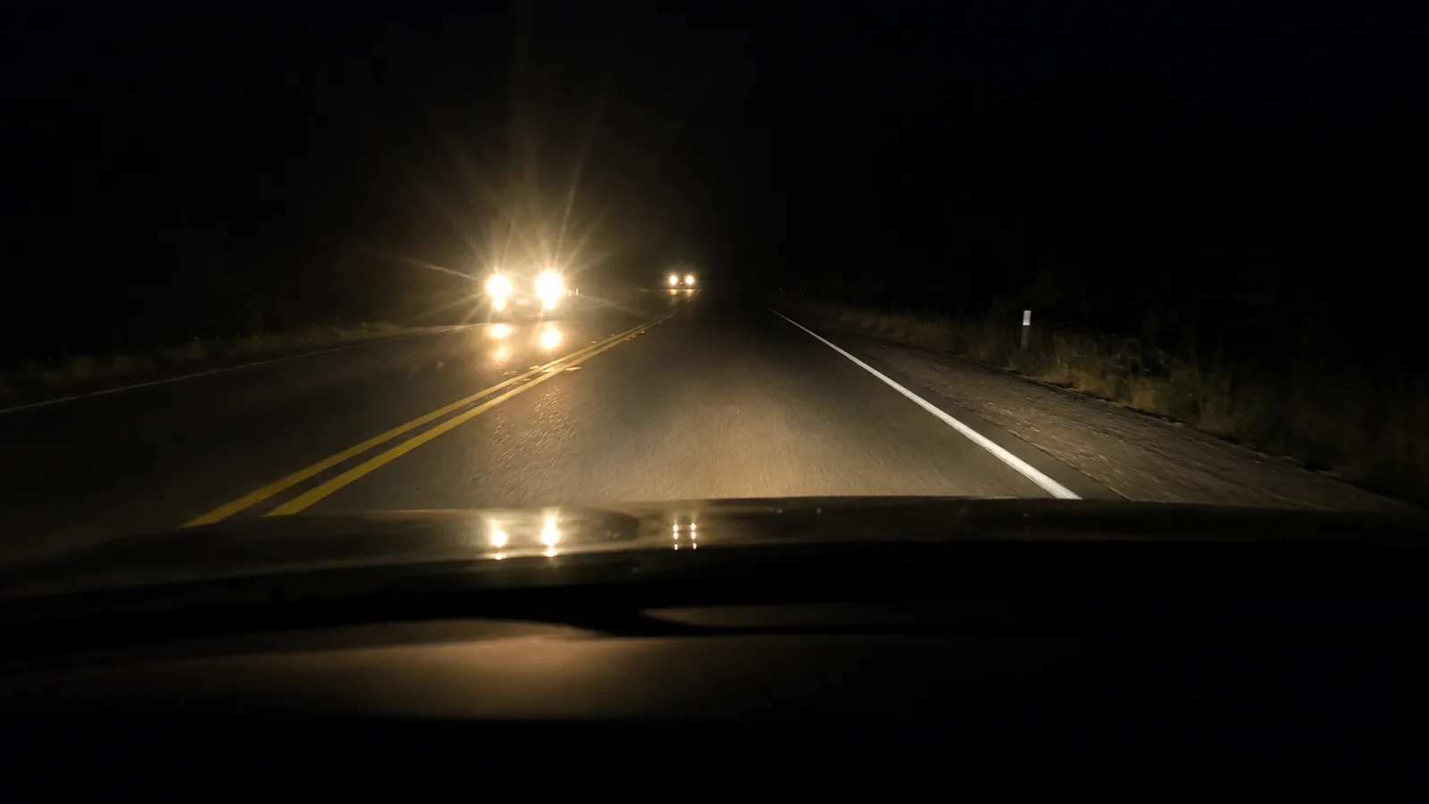 Night view from the driver's seat: an oncoming car in the far lane, its headlights glaring across a double yellow line.